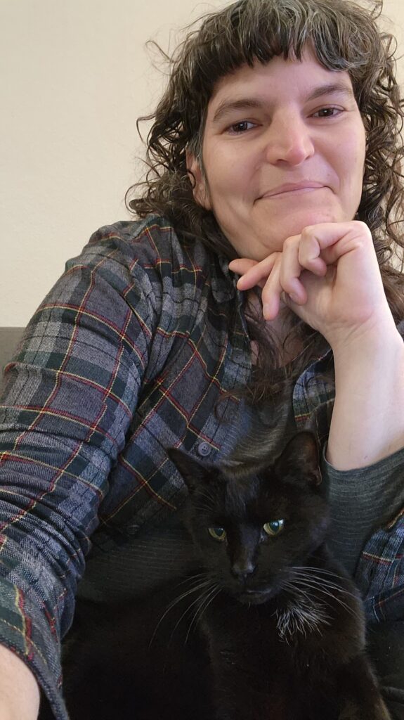 nicole gugliotti smiling in a selfie. She has short bangs and brown hair and is wearing a blue flannel shirt. She is posing with her black cat.