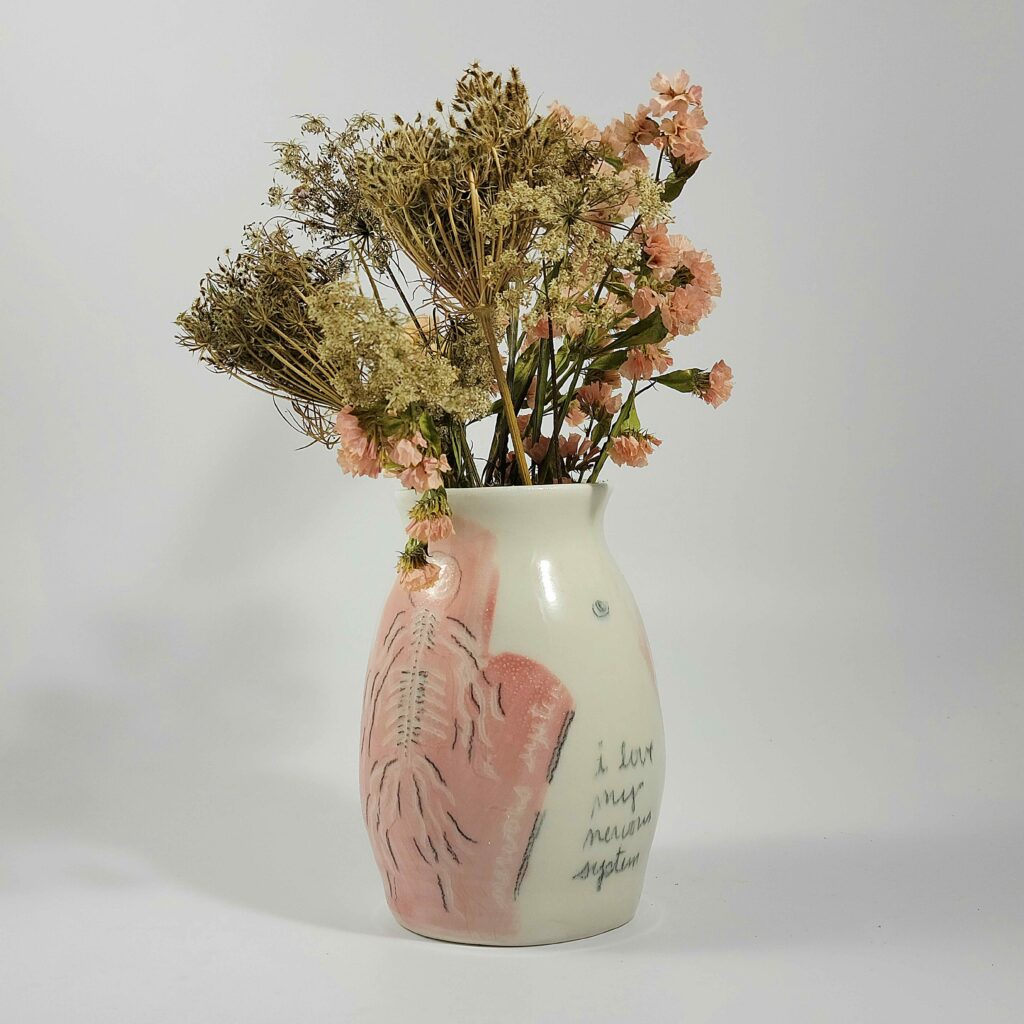 A pink and white vase with dried flowers created by nicole gugliotti. The vase depicts a human body with text that says "I love my nervous system".