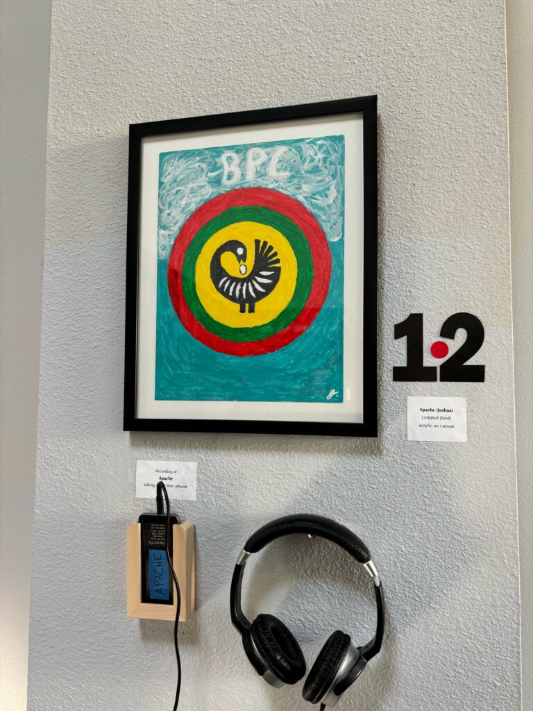 The photograph shows a painting of a stylized black Sankofa bird within a yellow, then green and finally red circle. The background of the painting is two shades of light blue with BPC at the top. Outside of the painting hangs a headset for viewers of the artwork to listen to the artist's description of the work.