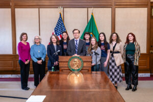 A group of activists with Governor Bob Ferguson in front of a row of flags